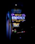 A vibrant neon sign displays the words 'HOLY SMOKE' along with the taglines 'T-SHIRTS', 'CIGARS', and 'VAPES'. The sign is predominantly blue, yellow, and purple, with stylized clouds accentuating the text. Below this sign, another illuminated sign advertises 'The Drunken Fry'. The surrounding area appears to be a bustling street with other signs and lights in the distance.