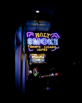 A vibrant neon sign displays the words 'HOLY SMOKE' along with the taglines 'T-SHIRTS', 'CIGARS', and 'VAPES'. The sign is predominantly blue, yellow, and purple, with stylized clouds accentuating the text. Below this sign, another illuminated sign advertises 'The Drunken Fry'. The surrounding area appears to be a bustling street with other signs and lights in the distance.