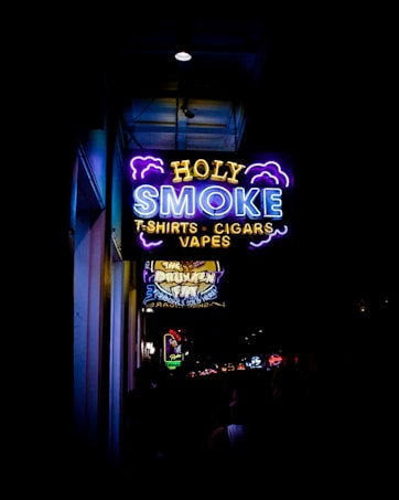 A vibrant neon sign displays the words 'HOLY SMOKE' along with the taglines 'T-SHIRTS', 'CIGARS', and 'VAPES'. The sign is predominantly blue, yellow, and purple, with stylized clouds accentuating the text. Below this sign, another illuminated sign advertises 'The Drunken Fry'. The surrounding area appears to be a bustling street with other signs and lights in the distance.