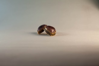 A pair of ornamental rings with large, dome-shaped gemstones. The gemstones are purple and set within intricate gold bands. The rings are placed on a light, neutral-colored surface with a gradient background.