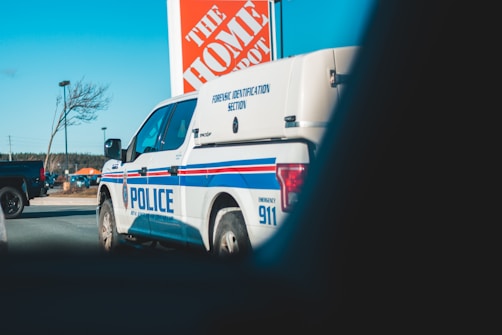 A police forensic identification vehicle parked near a Home Depot store. The background includes a clear sky, a tree, a streetlight, and another vehicle.