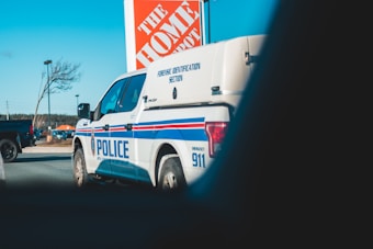 A police forensic identification vehicle parked near a Home Depot store. The background includes a clear sky, a tree, a streetlight, and another vehicle.