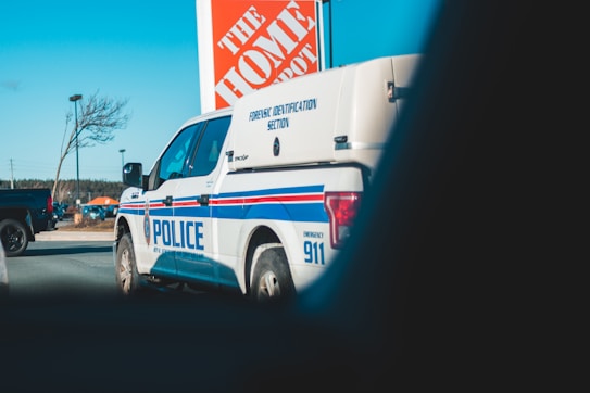 A police forensic identification vehicle parked near a Home Depot store. The background includes a clear sky, a tree, a streetlight, and another vehicle.