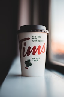 A takeaway coffee cup with black lid featuring festive holiday graphics, including a maple leaf and small red berries, sits on a windowsill. The text on the cup reads 'It's the most wonderful Tims of the year' in red and green letters.