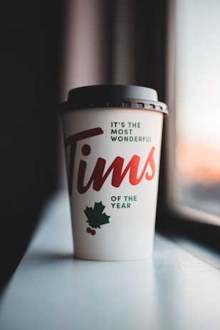 A takeaway coffee cup with black lid featuring festive holiday graphics, including a maple leaf and small red berries, sits on a windowsill. The text on the cup reads 'It's the most wonderful Tims of the year' in red and green letters.