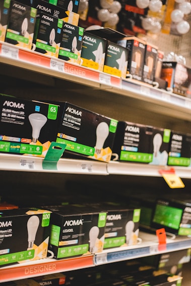 Shelves filled with various types of boxed LED light bulbs are visible, prominently featuring the NOMA brand. The boxes are black with green accents and display images of light bulbs and energy efficiency ratings. Price tags and product information are visible on the shelf edges.