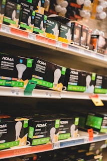 Shelves filled with various types of boxed LED light bulbs are visible, prominently featuring the NOMA brand. The boxes are black with green accents and display images of light bulbs and energy efficiency ratings. Price tags and product information are visible on the shelf edges.