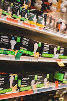 Shelves filled with various types of boxed LED light bulbs are visible, prominently featuring the NOMA brand. The boxes are black with green accents and display images of light bulbs and energy efficiency ratings. Price tags and product information are visible on the shelf edges.