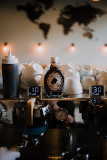 A cozy coffee shop ambiance with a bottle of syrup prominently placed among stacked white ceramic cups. Behind the cups, a metal espresso machine features digital displays showing numbers. Warm light bulbs illuminate the area with soft, ambient lighting, and there is a blurred map of the world on the wall, creating a welcoming atmosphere.