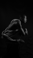A person is seated in a crouched position with a thick rope wrapped around their torso. The lighting creates dramatic shadows, highlighting the contours of their muscles. The monochrome palette adds to the starkness of the composition.