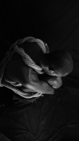 A person is tightly curled up into a fetal position, with thick rope wrapped around their shoulders and torso. The lighting is dramatic, casting strong shadows and highlighting the textures of the skin and rope. The overall composition is in black and white, creating a stark and intense visual impact.