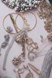 An assortment of jewelry including pearl-studded necklaces, large hoop earrings with inscriptions, gold chains, and various rings and bracelets arranged on a flat surface. Notable elements include the decorative pieces encrusted with rhinestones and intricate metalwork.