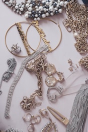 An assortment of jewelry including pearl-studded necklaces, large hoop earrings with inscriptions, gold chains, and various rings and bracelets arranged on a flat surface. Notable elements include the decorative pieces encrusted with rhinestones and intricate metalwork.