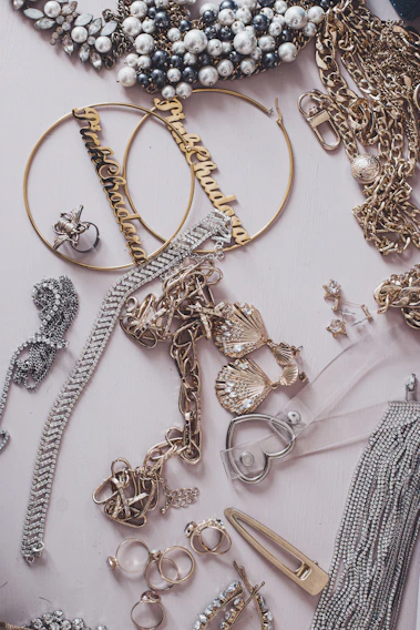 An assortment of jewelry including pearl-studded necklaces, large hoop earrings with inscriptions, gold chains, and various rings and bracelets arranged on a flat surface. Notable elements include the decorative pieces encrusted with rhinestones and intricate metalwork.