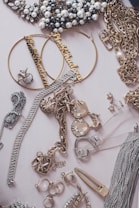An assortment of jewelry including pearl-studded necklaces, large hoop earrings with inscriptions, gold chains, and various rings and bracelets arranged on a flat surface. Notable elements include the decorative pieces encrusted with rhinestones and intricate metalwork.