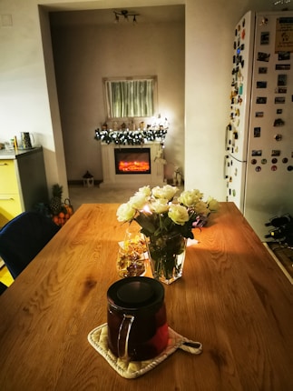 A cozy interior featuring a wooden dining table with a dark teapot on a mat, surrounded by a glass vase of white roses and a jar of wrapped candies. To the right, a refrigerator adorned with magnets stands. In the background, a decorative electric fireplace provides warmth, with a string of lights and figurines on the mantle. The setting is pleasantly lit, enhancing the intimate atmosphere.
