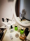 Various beauty and skincare products are neatly arranged on a white dressing table with a round mirror. The assortment includes perfumes, creams, and boxes, some with recognizable labels. Next to the mirror, a book sits with a decorative item resembling cotton branches in the background.