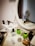 Various beauty and skincare products are neatly arranged on a white dressing table with a round mirror. The assortment includes perfumes, creams, and boxes, some with recognizable labels. Next to the mirror, a book sits with a decorative item resembling cotton branches in the background.