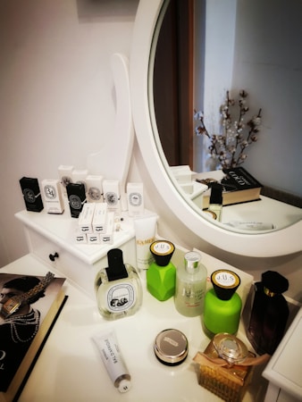 Various beauty and skincare products are neatly arranged on a white dressing table with a round mirror. The assortment includes perfumes, creams, and boxes, some with recognizable labels. Next to the mirror, a book sits with a decorative item resembling cotton branches in the background.
