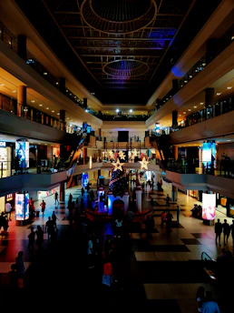 A multi-level shopping mall is bustling with people. The interior features large circular light fixtures on the ceiling, bright advertising screens, and a prominent central decoration with stars and an illuminated Christmas tree. The atmosphere is warm and inviting, with a combination of natural and artificial lighting highlighting different areas.