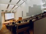 A modern lecture hall with tiered wooden seating, filled with students attentively watching a presentation on a large screen. The room is spacious with sleek architectural design, featuring high ceilings and white walls with geometric patterns. There is a lecturer standing near the podium delivering a lecture.