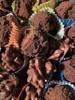 A variety of chocolate confections, including truffles dusted with cocoa powder and clusters of chocolate-covered nuts, are arranged together. The sweets are presented in colorful paper cups, adding a visually appealing contrast.