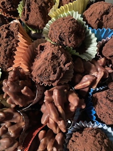A variety of chocolate confections, including truffles dusted with cocoa powder and clusters of chocolate-covered nuts, are arranged together. The sweets are presented in colorful paper cups, adding a visually appealing contrast.