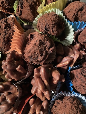 A variety of chocolate confections, including truffles dusted with cocoa powder and clusters of chocolate-covered nuts, are arranged together. The sweets are presented in colorful paper cups, adding a visually appealing contrast.