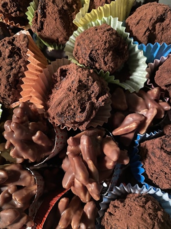 A variety of chocolate confections, including truffles dusted with cocoa powder and clusters of chocolate-covered nuts, are arranged together. The sweets are presented in colorful paper cups, adding a visually appealing contrast.