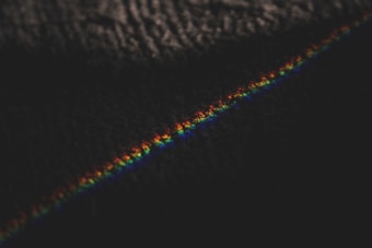 A dark background is intersected by a thin, colorful line displaying the spectrum of a rainbow. The line appears slightly textured and is composed of colors ranging from red to violet.