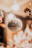 A decorative Christmas ornament featuring a black and white sketch of Wroclaw's architecture hangs in focus, against a blurred background filled with warm lights. Nearby, a miniature car ornament can be seen partially.