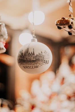 A decorative Christmas ornament featuring a black and white sketch of Wroclaw's architecture hangs in focus, against a blurred background filled with warm lights. Nearby, a miniature car ornament can be seen partially.