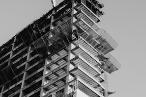 A black and white photograph capturing the upward perspective of a high-rise building under construction. The structural framework of the building is prominently visible with scaffolding and protective netting in place around the edges. The image highlights the intricate details of the construction process and the geometric patterns formed by the unfinished floors.