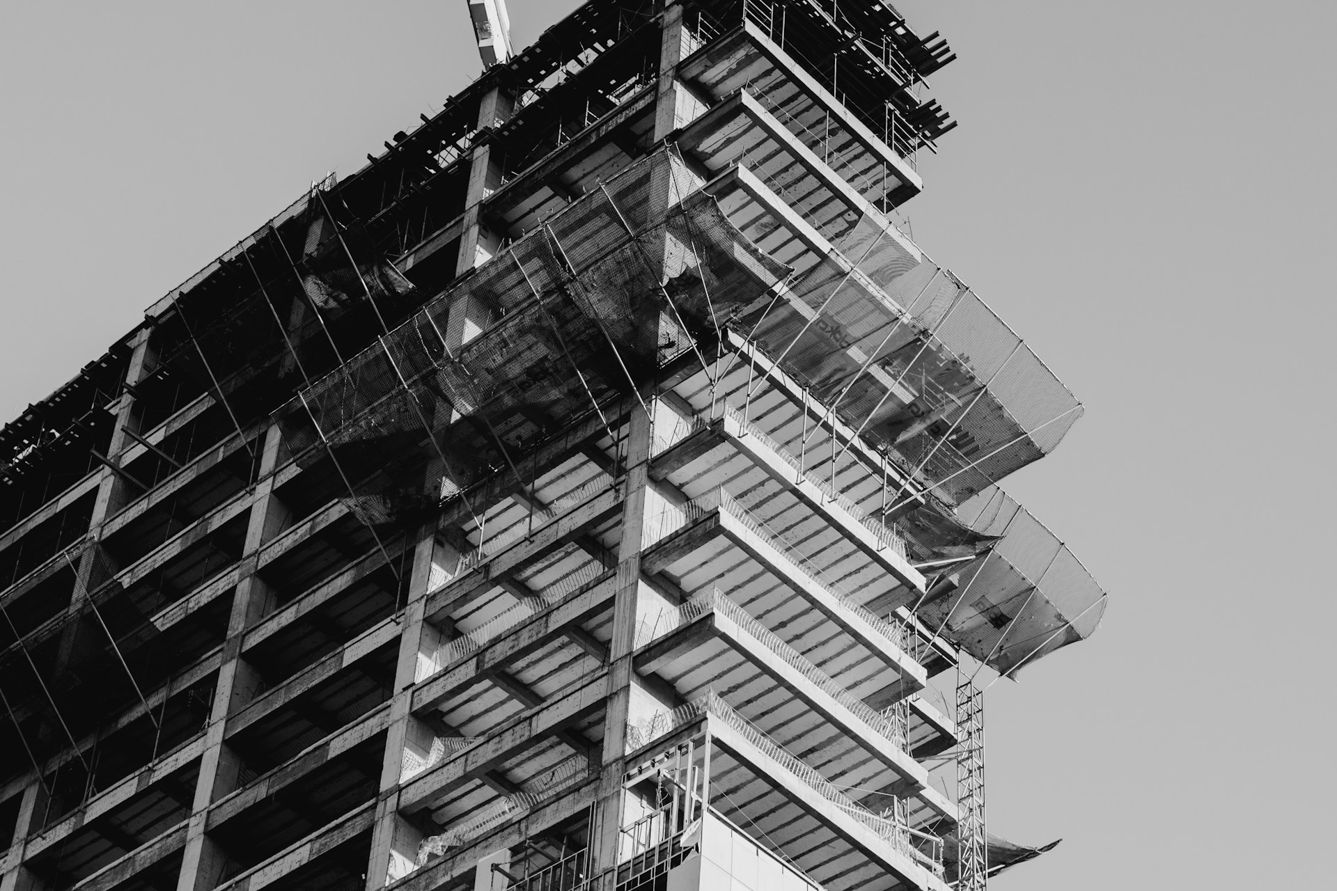 A black and white photograph capturing the upward perspective of a high-rise building under construction. The structural framework of the building is prominently visible with scaffolding and protective netting in place around the edges. The image highlights the intricate details of the construction process and the geometric patterns formed by the unfinished floors.