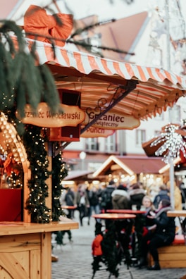 A festive outdoor market scene with wooden stalls decorated with lights and garlands. There are people walking and gathering around tables. One of the stalls has a large red bow on top and signs with text in a foreign language. A Christmas-like atmosphere is present with elements of holiday decoration and subtle lighting.