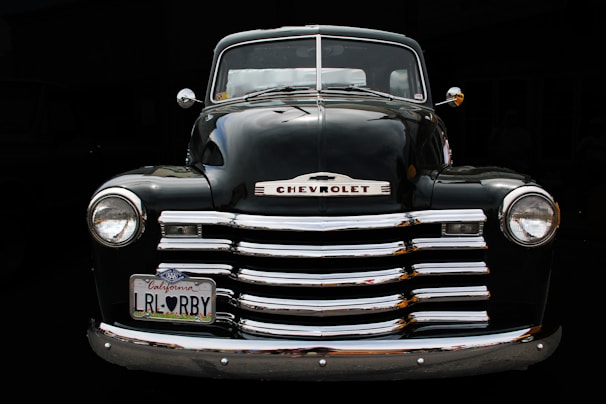 A vintage Chevrolet truck with a shiny chrome grille and a distinctively large front bumper. The vehicle features round headlights and has a license plate from California. The truck's body is a sleek black color, reflecting surrounding light.