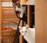 Two cats are situated in an indoor setting. A multicolored cat is perched on a chair, peering inquisitively, while a white cat stands on the floor in front. In the background, a wooden basket filled with orange fabric is visible, along with a dining arrangement featuring chairs and a table topped with a cloth.