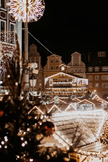 Festive city square scene at night with bright Christmas lights illuminating the area. Traditional European architecture forms the backdrop, adorned with decorative garlands and light displays. A large star-shaped decoration is suspended above, while a Christmas tree with ornaments is visible in the foreground.