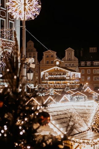 Festive city square scene at night with bright Christmas lights illuminating the area. Traditional European architecture forms the backdrop, adorned with decorative garlands and light displays. A large star-shaped decoration is suspended above, while a Christmas tree with ornaments is visible in the foreground.