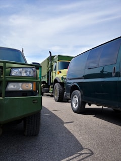 Several vehicles including a military-style truck and a van are parked closely together on a paved road. The dominant green tones of the vehicles contrast with the clear blue sky overhead.