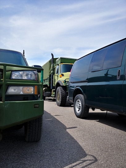 Several vehicles including a military-style truck and a van are parked closely together on a paved road. The dominant green tones of the vehicles contrast with the clear blue sky overhead.