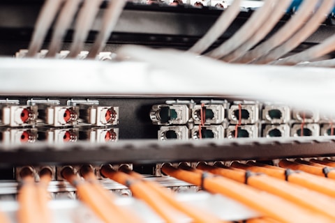 A close-up view of a network server or data center with numerous cables and connections. Orange cables are neatly arranged in the foreground, while various ports and connectors are visible in the background.