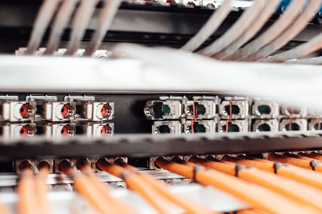 A close-up view of a network server or data center with numerous cables and connections. Orange cables are neatly arranged in the foreground, while various ports and connectors are visible in the background.
