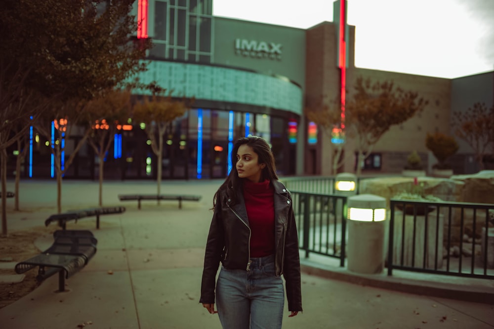 woman in black leather jacket and blue denim jeans standing on sidewalk during night time
