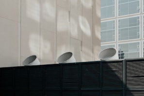 Three industrial ventilation units protruding from a dark metal fence are set against a light-colored building with rectangular window frames. The image features a contrast between the dark, horizontal slats of the fence and the smooth, vertical lines of the building alongside the geometric patterns of light and shadow on the wall.