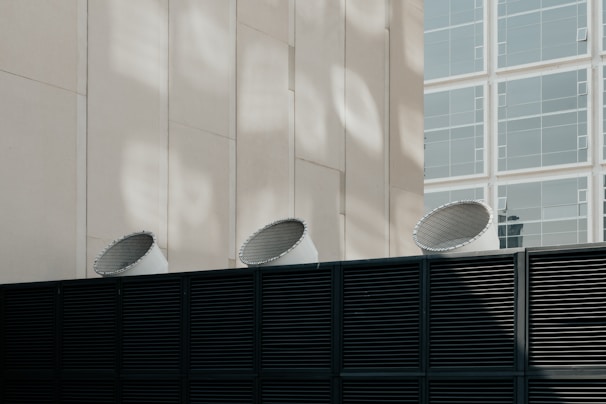 Three industrial ventilation units protruding from a dark metal fence are set against a light-colored building with rectangular window frames. The image features a contrast between the dark, horizontal slats of the fence and the smooth, vertical lines of the building alongside the geometric patterns of light and shadow on the wall.