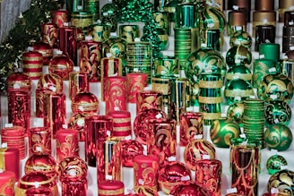 A diverse collection of decorated candles is arranged on a table. The candles display intricate designs in red and green with gold accents, creating a festive appearance. Some candles are cylindrical while others are round, resembling ornaments. The overall arrangement suggests a holiday or Christmas theme.