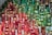 A diverse collection of decorated candles is arranged on a table. The candles display intricate designs in red and green with gold accents, creating a festive appearance. Some candles are cylindrical while others are round, resembling ornaments. The overall arrangement suggests a holiday or Christmas theme.