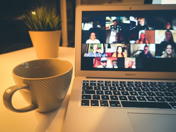 A cozy setup with a laptop displaying a video conference with multiple participants. In the foreground, there is a large mug on the desk next to the laptop. A small potted plant is in the background, adding a touch of greenery to the scene. The lighting is warm, creating a comfortable atmosphere for remote communication.