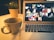 A cozy setup with a laptop displaying a video conference with multiple participants. In the foreground, there is a large mug on the desk next to the laptop. A small potted plant is in the background, adding a touch of greenery to the scene. The lighting is warm, creating a comfortable atmosphere for remote communication.